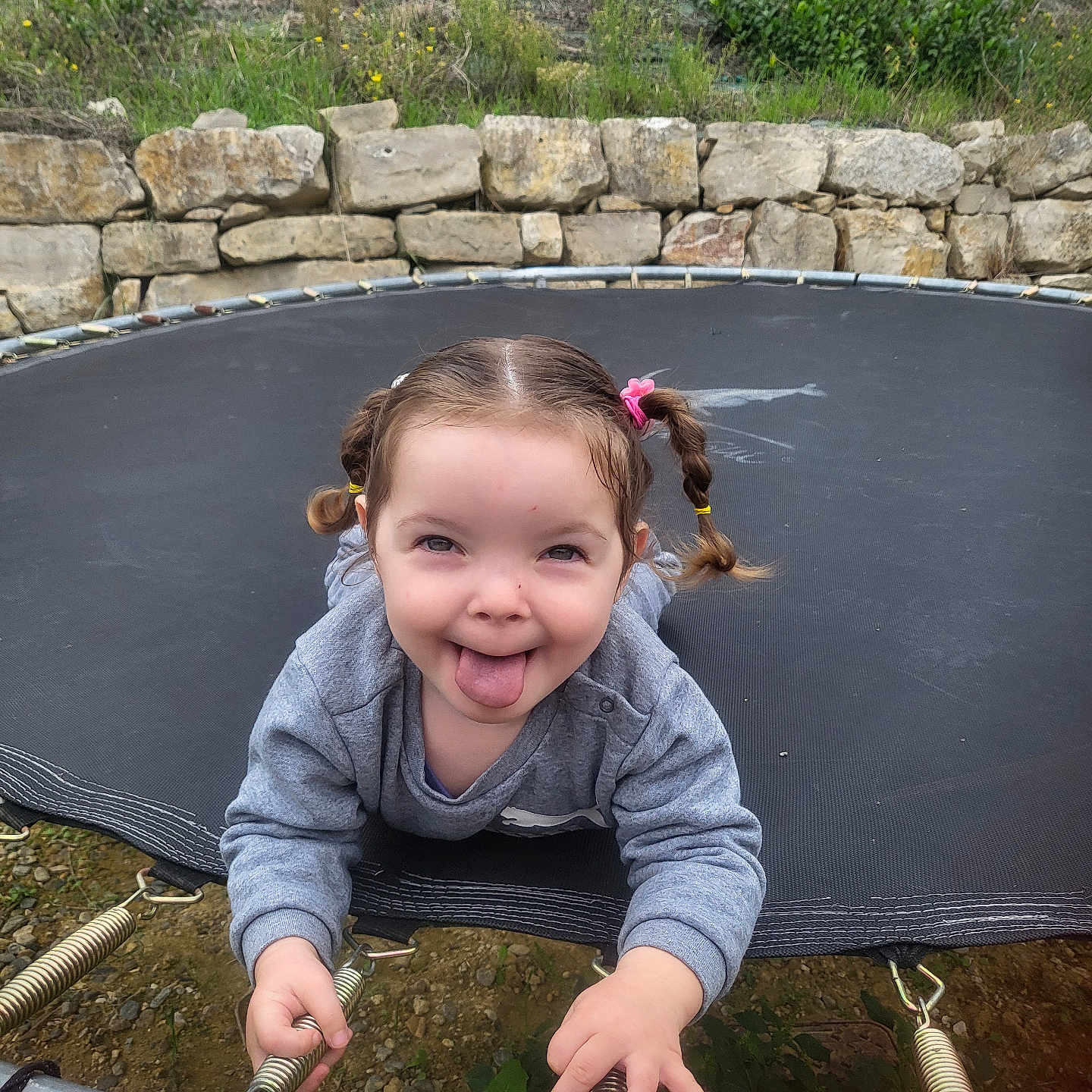 Ana Matilda a rejoint le concours — aidez-le/la à gagner de superbes lots ! casual_clothing, child, expression, fun, grass, gray_sweatshirt, happy, holding, nature, outdoor, person, pigtails, playful, smiling, springs, stone_wall, tongue, tongue_out, trampoline, young_child