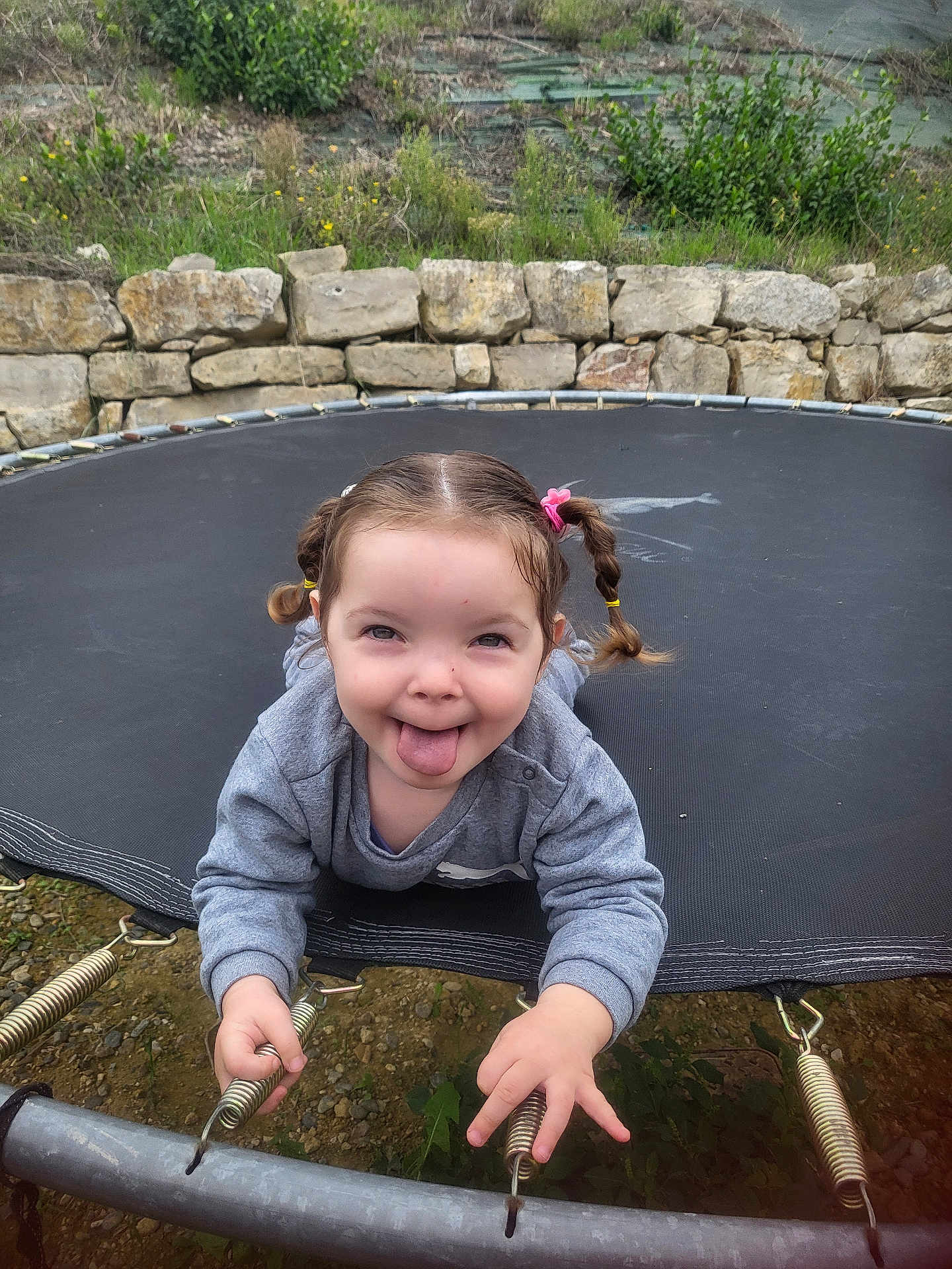 Ana Matilda a rejoint le concours — aidez-le/la à gagner de superbes lots ! child, pigtails, tongue_out, trampoline, playful, springs, gray_sweatshirt, outdoor, grass, stone_wall, smiling, happy, young_child, person, fun, nature, expression, tongue, holding, casual_clothing