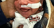 Anthony is registered to the contest to win money with this photo: newborn, baby, smile, blanket, hand, face, medical_tape, clothing, infant, person, cute, sleepy, resting, wrapped, cozy, warm, skin, head, portrait, child