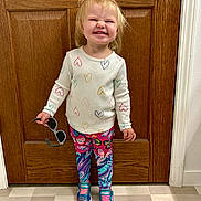 Riley is registered to the contest to win money with this photo: blonde_hair, butterfly_pattern, child, colorful_clothing, cute, door, floor, happy, heart_pattern, holding_sunglasses, indoor, leggings, long_sleeves, playful, portrait, smiling, sneakers, standing, toddler, wooden_door