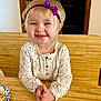 blonde_hair, child, clothing, cute, face, food, happy, headband, home, indoor, long_sleeve_shirt, person, plate, purple_bow, sitting, smiling, table, toddler, wooden_table, young_child