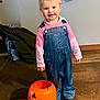 Riley is registered to the contest to win money with this photo: casual_clothing, child, cute, decor, denim_overalls, halloween, happy, headband, holiday, indoor, person, pink_hoodie, pumpkin_bucket, shoes, smiling, standing, toddler, toy, wall, wooden_floor