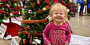 Riley is registered to the contest to win money with this photo: child, christmas_tree, clothing, event, family, festive, floor, gold_decor, happy, holiday_decorations, indoor, market, ornaments, people, person, red_ribbon, shopping, smiling, sneakers, toddler