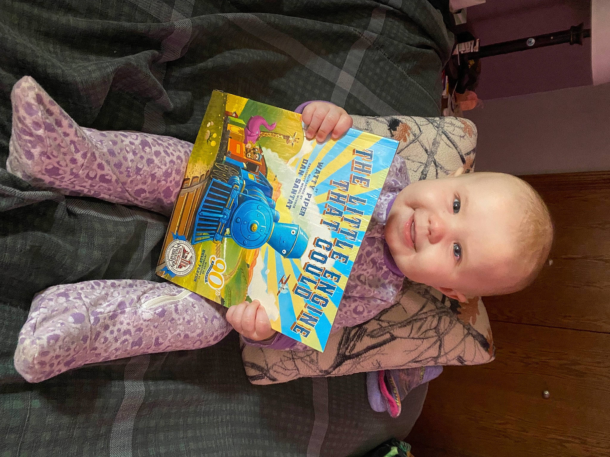 Riley is registered to the contest to win money with this photo: baby, baby_toddler_clothing, book, child, comfort, finger, happy, linens, person, product, publication, reading, room, sitting, smile, toddler
