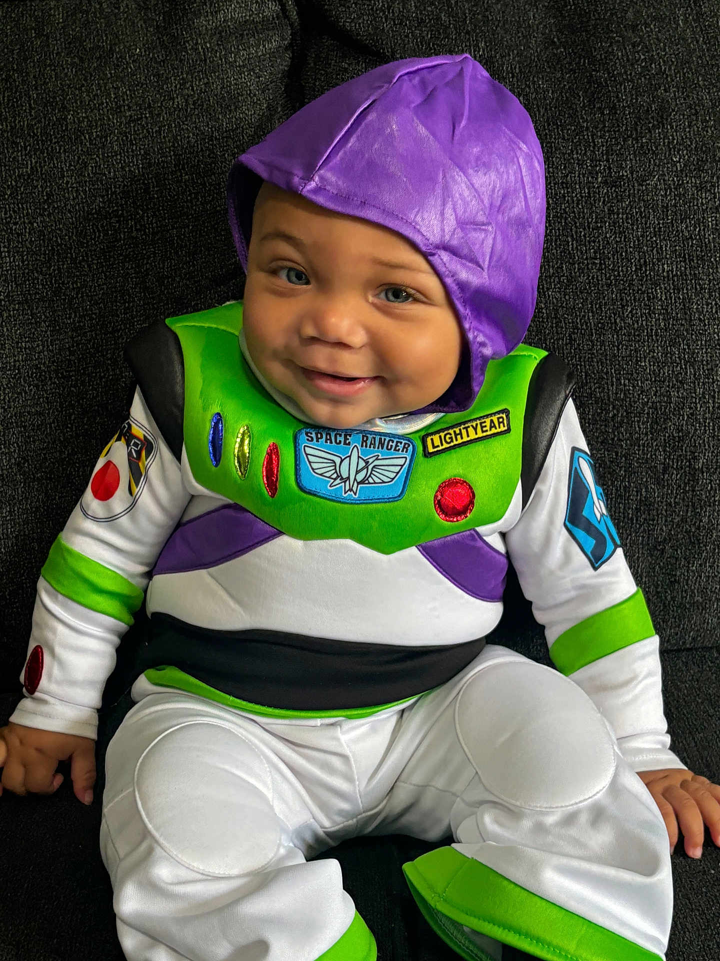 Silas is registered to the contest to win money with this photo: baby, child, costume, buzz_lightyear, purple_hood, smile, face, sitting, couch, clothing, green, white, black, indoor, cute, portrait, happy, infant, person, young