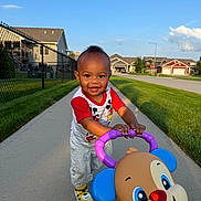 Micah is registered to the contest to win money with this photo: toddler, child, smiling, outdoor, sidewalk, toy, walker, puppy, grass, houses, sunlight, happy, person, footwear, clothing, red_shirt, blue_sky, fence, residential_area, playing