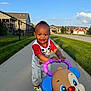 toddler, child, smiling, outdoor, sidewalk, toy, walker, puppy, grass, houses, sunlight, happy, person, footwear, clothing, red_shirt, blue_sky, fence, residential_area, playing