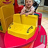 amusement, boat, child, face, floor_tiles, fun, hair, hands, happy, indoor, kiddie_ride, person, pink, playful, smiling, striped_shirt, stroller, toddler, toy, yellow