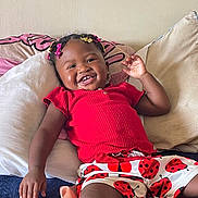 Xena is registered to the contest to win money with this photo: toddler, child, smiling, happy, indoor, pillow, bed, clothing, shorts, red, ladybug, hair_accessories, person, cute, portrait, skin, hand, relaxing, cushion, home