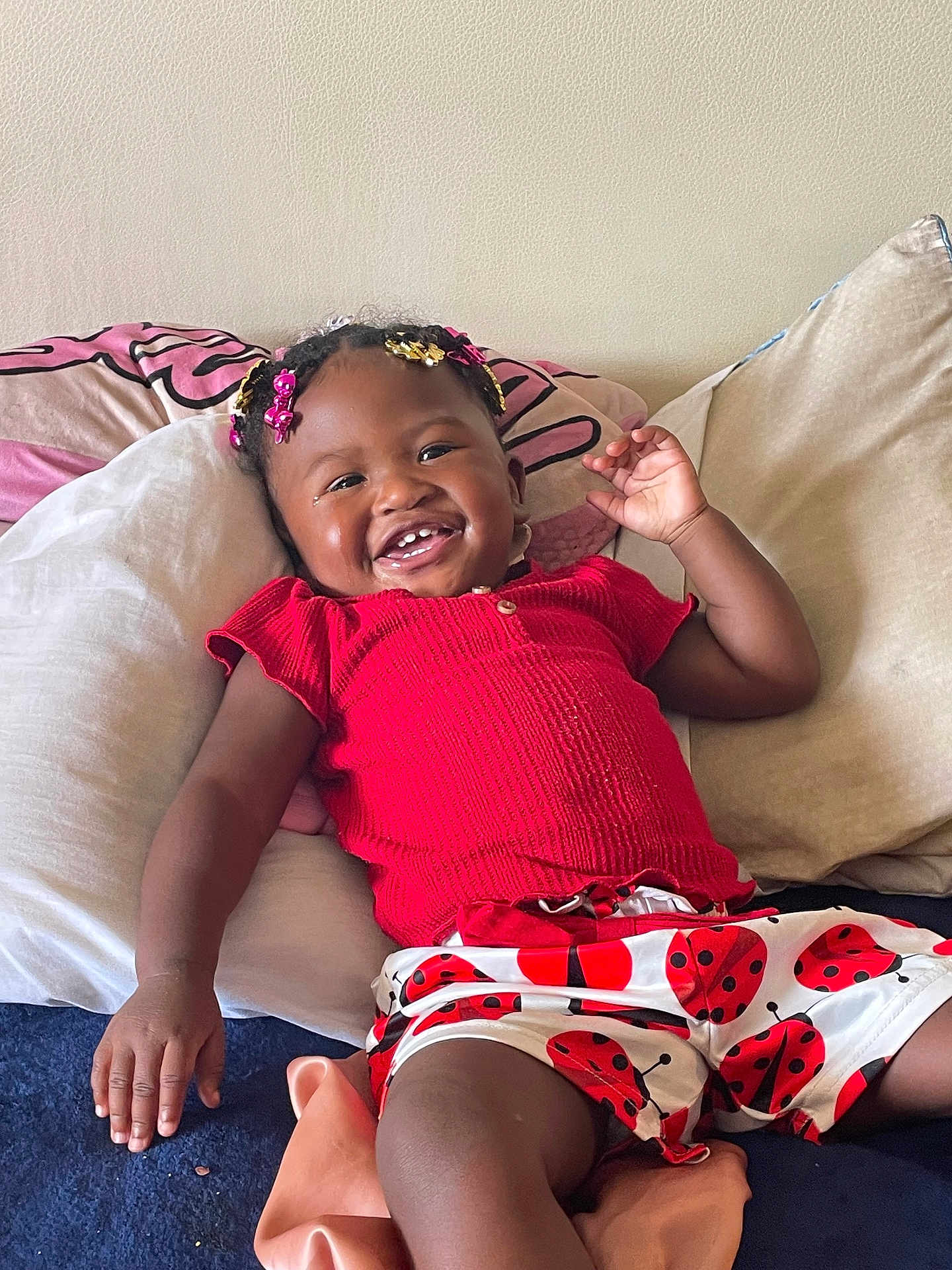 Xena is registered to the contest to win money with this photo: toddler, child, smiling, happy, indoor, pillow, bed, clothing, shorts, red, ladybug, hair_accessories, person, cute, portrait, skin, hand, relaxing, cushion, home