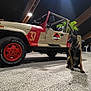 dog, jeep, jurassic_park, vehicle, indoor, garage, tire, red, beige, leash, pet, car, floor, plant, lighting, shadow, open_top, animal, sitting, black