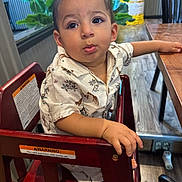 Enzo is registered to the contest to win money with this photo: toddler, child, high_chair, shirt, giraffe_pattern, wooden_chair, table, indoor, restaurant, plant, floor, foot, hand, face, curious, expression, furniture, warning_label, window, light