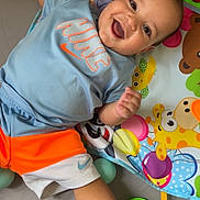 Kassim a rejoint le concours — aidez-le/la à gagner de superbes lots ! baby, smiling, blue_shirt, orange_shorts, nike_logo, play_mat, plush_toy, colorful_balls, happy, child, indoor, toy, foot, hand, face, clothing, infant, cute, lying_down, playtime