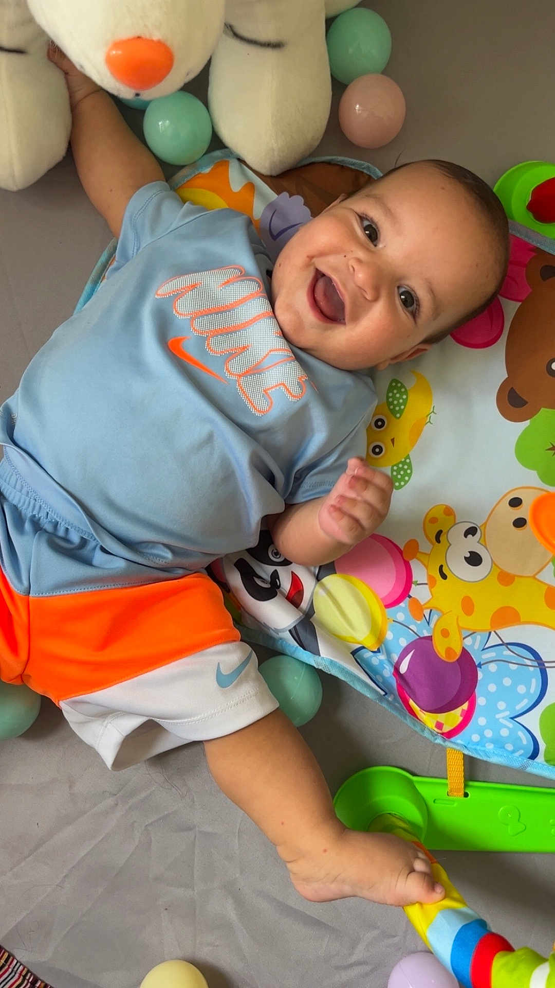 Kassim a rejoint le concours — aidez-le/la à gagner de superbes lots ! baby, smiling, blue_shirt, orange_shorts, nike_logo, play_mat, plush_toy, colorful_balls, happy, child, indoor, toy, foot, hand, face, clothing, infant, cute, lying_down, playtime