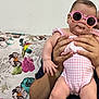 baby, pink_sunglasses, pink_outfit, person, adult_hands, floral_couch, indoor, cute, child, infant, fashion, accessories, sitting, portrait, home, furniture, casual, funny_glasses, young_child, holding