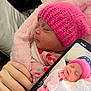 baby, newborn, pink_hat, blanket, sleeping, infant, hand, smartphone, screen, portrait, cozy, soft_texture, cute, child, clothing, knit_hat, wrapped, resting, indoor, person