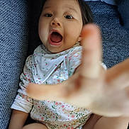 Jeanne a rejoint le concours — aidez-le/la à gagner de superbes lots ! baby, child, hand, reaching, floral_clothing, bib, open_mouth, couch, blue_fabric, indoor, cute, young_child, portrait, expression, playful, sitting, person, infant, cozy, blurred_hand