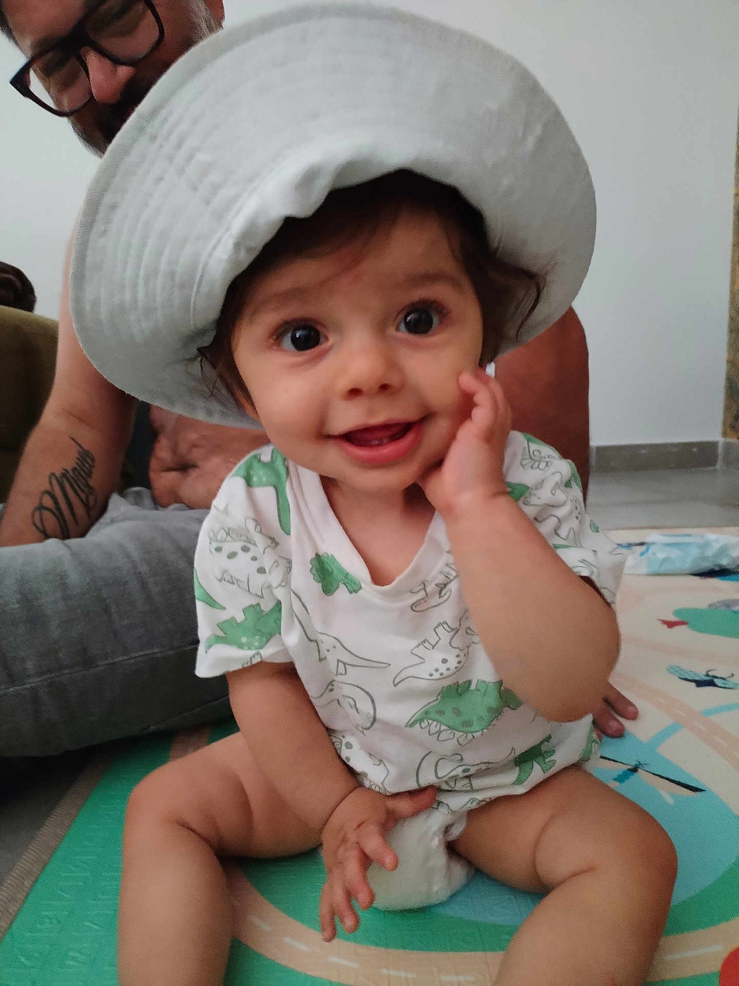 Joseph participe au concours pour gagner de l'argent avec cette photo : baby, child, hat, smile, dinosaur_print, tshirt, play_mat, indoor, adult, glasses, tattoo, happy, sitting, hand_on_face, floor, person, cute, family, joyful, portrait
