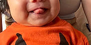 Skyler is registered to the contest to win money with this photo: baby, child, infant, orange_shirt, pumpkin_face, tongue_out, smiling, person, cute, happy, indoors, adult, white_tank_top, face, hair, skin, hand, clothing, holding, closeup