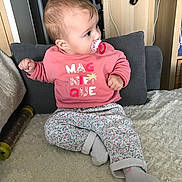 Lucie a rejoint le concours — aidez-le/la à gagner de superbes lots ! baby, pacifier, pink_sweatshirt, floral_pants, socks, cushion, blanket, indoor, child, furniture, home, curious, seated, cross_legged, person, young_child, soft_texture, casual_clothing, looking_away, cozy