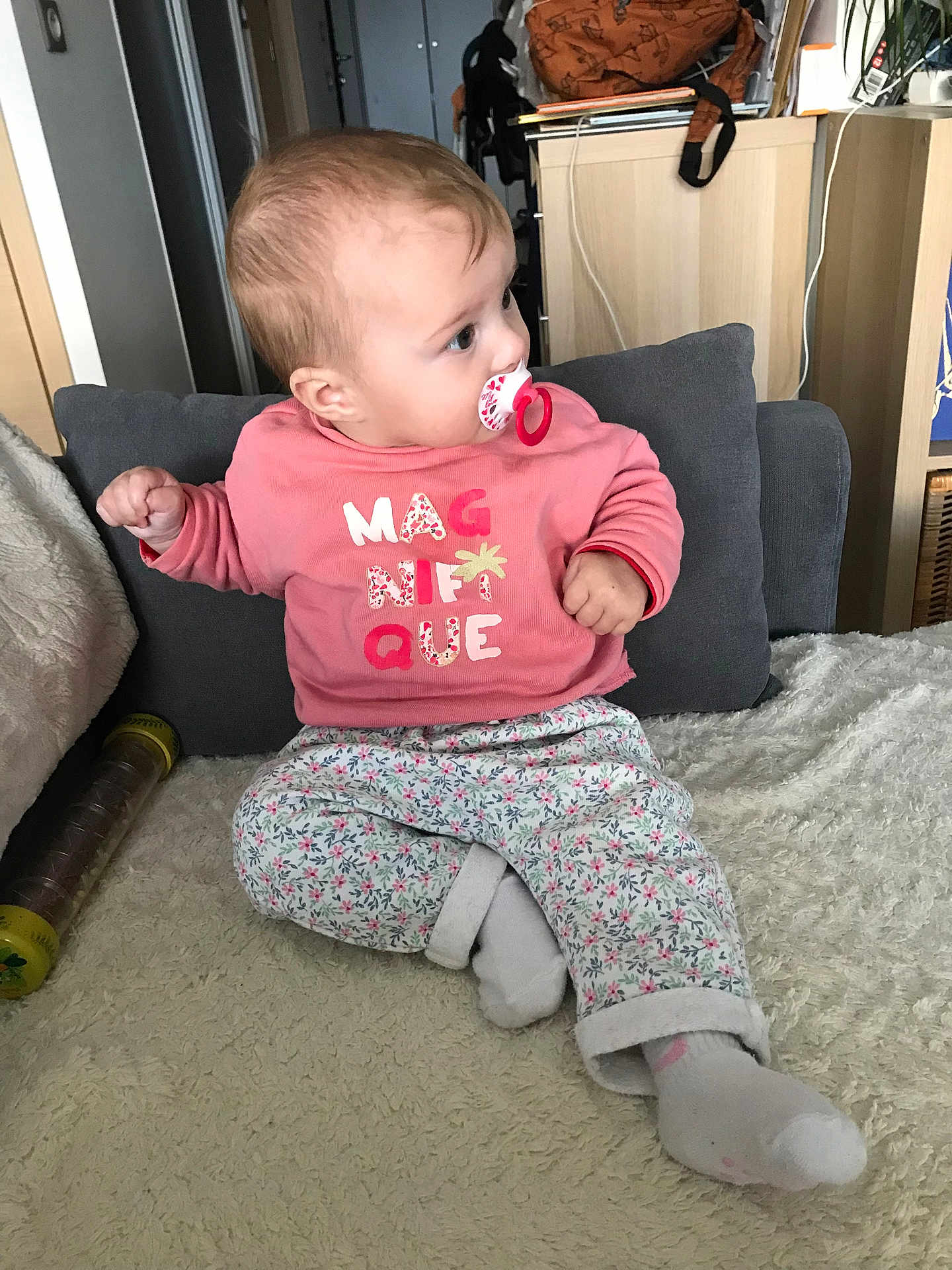 Lucie a rejoint le concours — aidez-le/la à gagner de superbes lots ! baby, pacifier, pink_sweatshirt, floral_pants, socks, cushion, blanket, indoor, child, furniture, home, curious, seated, cross_legged, person, young_child, soft_texture, casual_clothing, looking_away, cozy