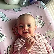 Grey is registered to the contest to win money with this photo: baby, infant, smiling, pink_clothing, blanket, elephant_pattern, cute, happy, face, child, indoors, natural_light, portrait, soft_texture, hand, young_child, cozy, nursery, sleeping_mat, adorable