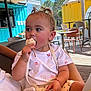 child, toddler, eating, ice_cream, outdoor, cafe, umbrella, table, chair, sunlight, summer, cute, hair_buns, white_clothing, bracelet, purse, pink_flower, earring, wooden_deck, relaxed