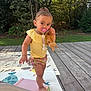 toddler, child, outdoor, play_mat, pacifier, plush_toy, yellow_top, floral_shorts, barefoot, grass, trees, wooden_deck, summer, cute, person, young_child, sunlight, nature, play, standing