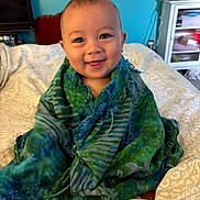 Kayu is registered to the contest to win money with this photo: baby, child, smiling, green_shawl, blue_shawl, bedspread, indoor, home, furniture, mirror, cabinet, television, portrait, cute, happy, person, sitting, blanket, cozy, wrapped