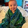 baby, child, smiling, green_shawl, blue_shawl, bedspread, indoor, home, furniture, mirror, cabinet, television, portrait, cute, happy, person, sitting, blanket, cozy, wrapped