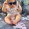 baby, child, sunglasses, hat, striped_clothing, romper, sitting, couch, floral_pattern, outdoor, sunlight, cute, chubby, foot, hand, face, portrait, summer, shade, relaxation