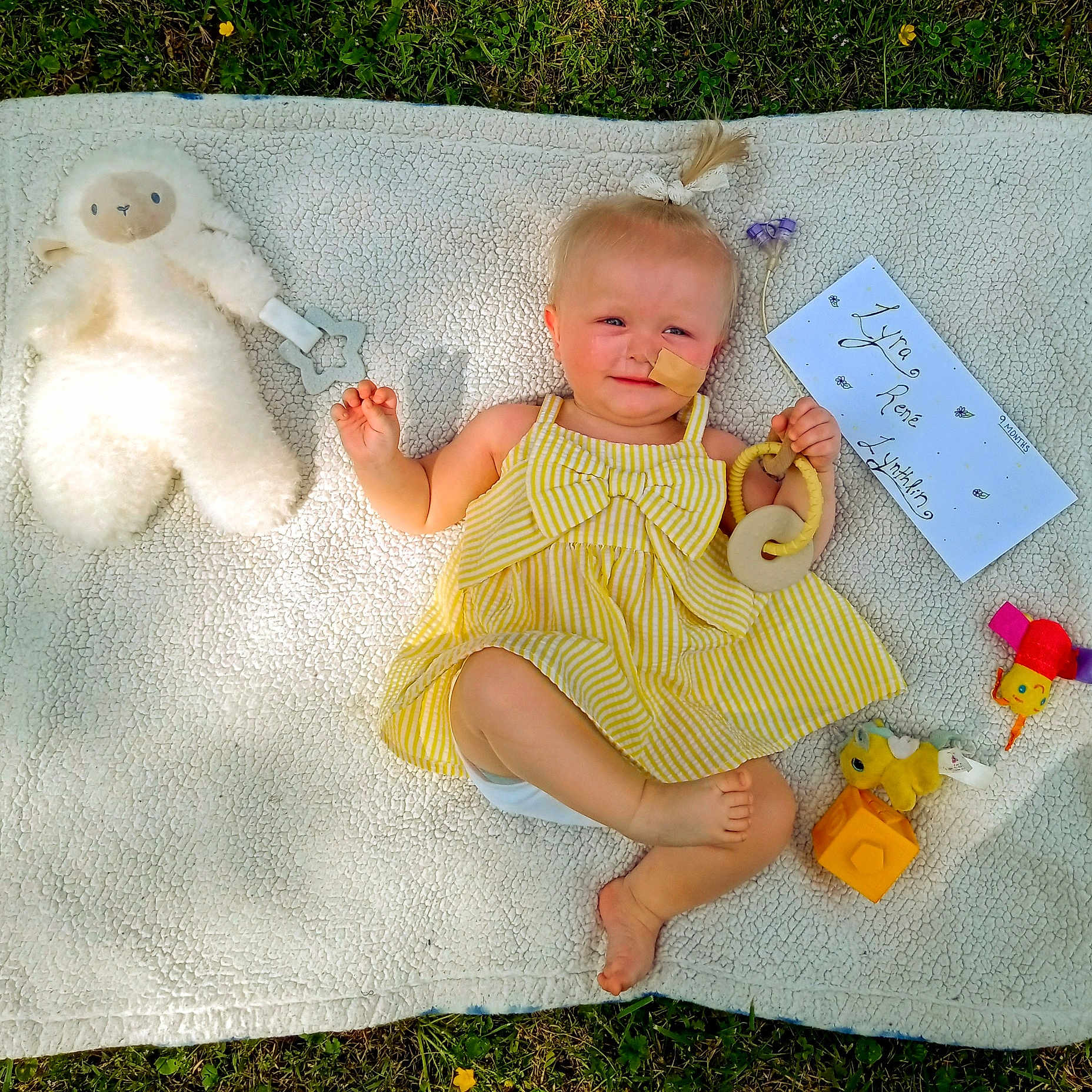 Lyra joined the competition — help win amazing prizes! baby, bandage, blanket, child, cute, dress, face, grass, grass_background, handwritten_note, happy, infant, outdoor, person, playing, smile, soft_texture, sunlight, toy, yellow