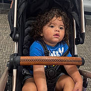 Carlo joined the competition — help win amazing prizes! toddler, child, stroller, curly_hair, blue_shirt, black_socks, sitting, portrait, indoors, airport, waiting_room, seated, baby_carriage, harness, curious, face, eyes, carpet, chairs, travel