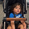 toddler, child, stroller, curly_hair, blue_shirt, black_socks, sitting, portrait, indoors, airport, waiting_room, seated, baby_carriage, harness, curious, face, eyes, carpet, chairs, travel