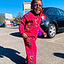 child, smiling, outdoor, parking_lot, car, glasses, red_clothing, knitwear, flowers, shoes, sunny, shadow, asphalt, person, happy, walking, hair_accessories, urban, daytime, cute