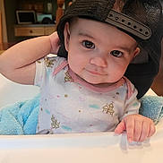 Olivia is registered to the contest to win money with this photo: baby, child, high_chair, black_cap, mesh_cap, curious_look, indoor, kitchen, blue_blanket, infant, cute, portrait, casual_clothing, hand, face, person, toddlers, smiling, seated, home