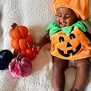 Genevieve joined the competition — help win amazing prizes! baby, child, pumpkin_costume, orange, green, smiling, happy, sitting, blanket, decorative_pumpkins, plush, cute, holiday, autumn, festive, indoors, soft_texture, infant, joyful, portrait