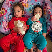 Marcello is registered to the contest to win money with this photo: children, toddlers, siblings, babies, bottles, feeding, seated, chair, colorful, floral, princess, cartoon, clothing, red_outfit, blue_outfit, indoors, portrait, faces, family, cute