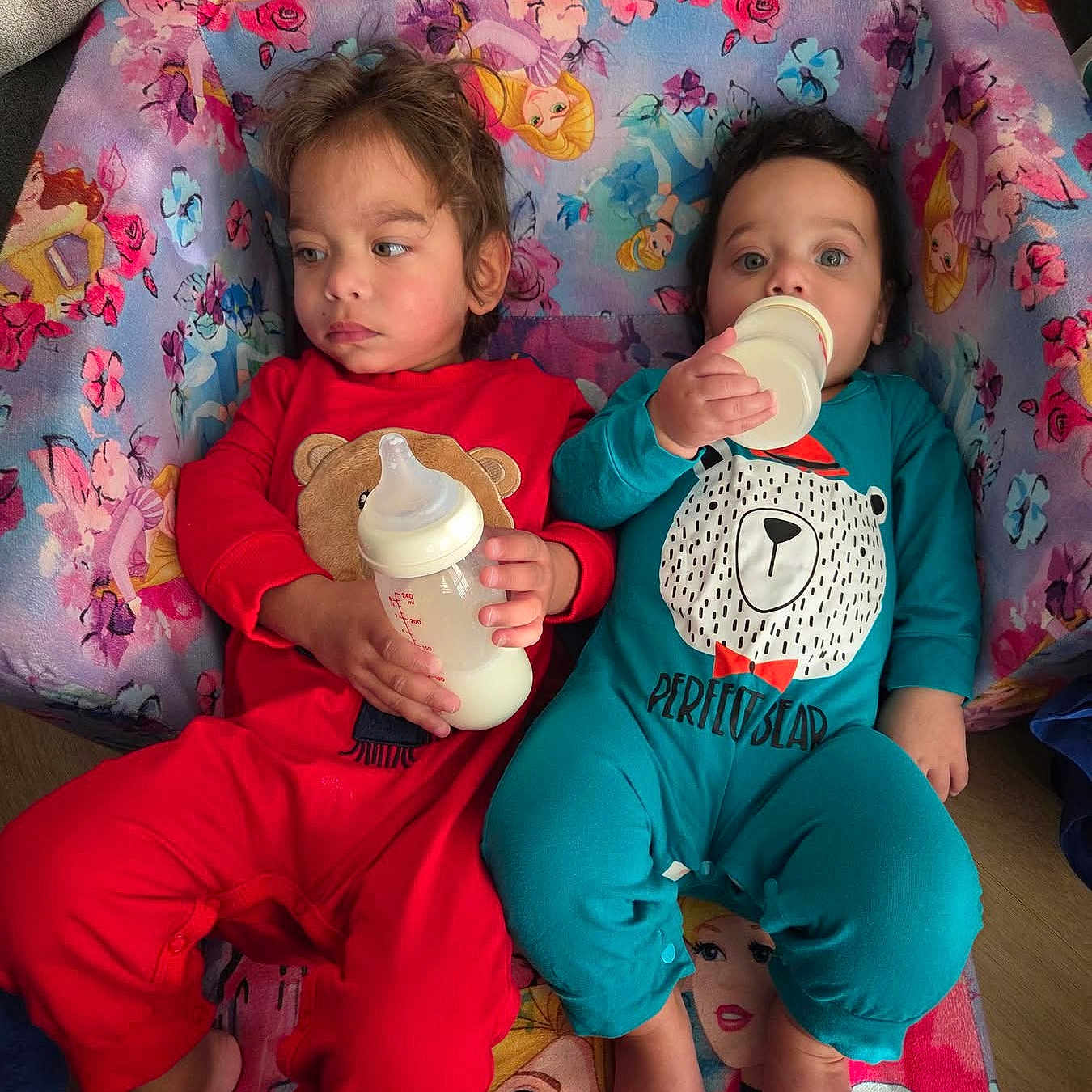 Marcello is registered to the contest to win money with this photo: children, toddlers, siblings, babies, bottles, feeding, seated, chair, colorful, floral, princess, cartoon, clothing, red_outfit, blue_outfit, indoors, portrait, faces, family, cute