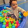 toddler, child, ball_pit, colorful_balls, blue_balls, yellow_balls, pink_balls, green_balls, red_balls, knitted_sweater, patterned_pants, curly_hair, indoor, floor, play, happy, smiling, plastic_container, cute, joyful