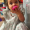 Leearnah is registered to the contest to win money with this photo: child, toddler, girl, portrait, indoors, sitting, dress, floral_dress, curly_hair, brown_hair, brown_eyes, smiling, curious, toy, teether, hand, necklace, playroom, houseplant, wooden_floor