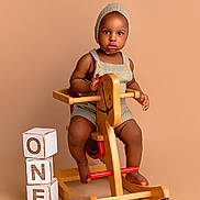 Eze is registered to the contest to win money with this photo: baby, blocks, brown_skin, child, floor, footwear_none, furniture, indoor, isolated_background, knitted_bonnet, one, play, portrait, rocking_horse, romper, serious_expression, studio, toddler, toy, wooden_toy