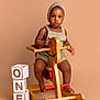 baby, blocks, brown_skin, child, floor, footwear_none, furniture, indoor, isolated_background, knitted_bonnet, one, play, portrait, rocking_horse, romper, serious_expression, studio, toddler, toy, wooden_toy