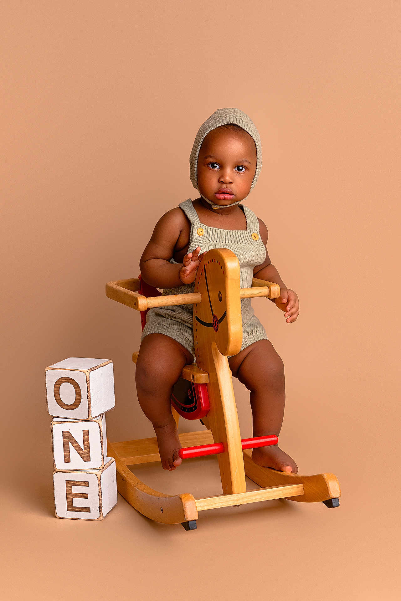 Eze is registered to the contest to win money with this photo: baby, blocks, brown_skin, child, floor, footwear_none, furniture, indoor, isolated_background, knitted_bonnet, one, play, portrait, rocking_horse, romper, serious_expression, studio, toddler, toy, wooden_toy