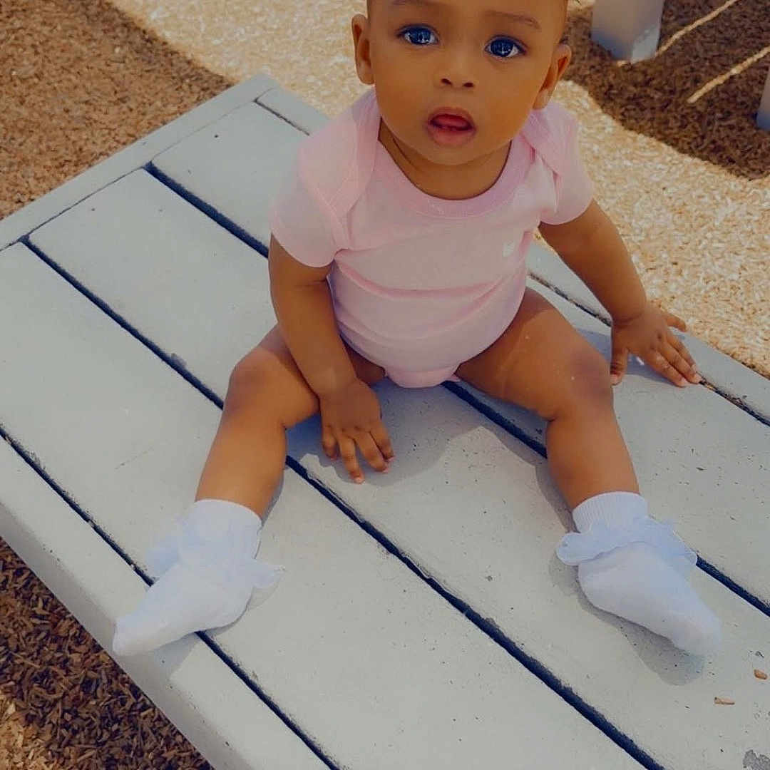 Rhayn joined the competition — help win amazing prizes! baby, bench, brick_wall, child, curly_hair, cute, expression, face, hands, infant, legs, outdoor, person, pink_onesie, playground, sitting, socks, sunlight, wood_chips, young_child
