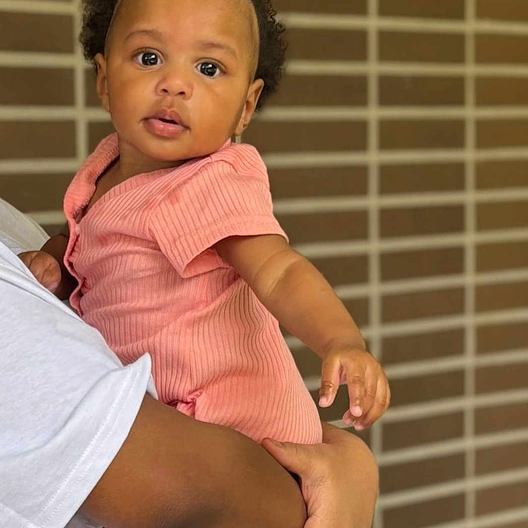 Khaïlys a rejoint le concours — aidez-le/la à gagner de superbes lots ! arm, baby, background, brick_wall, child, curly_hair, cute, expression, face, foot, hand, holding, indoor, looking, person, pink_clothing, portrait, skin, toddler, topknot