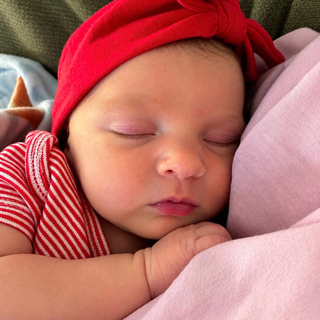 Aria is registered to the contest to win money with this photo: newborn, baby, sleeping, portrait, face, red_hat, striped_clothing, pink_clothing, infant, closed_eyes, peaceful, cuddling, indoors, close_up, soft_lighting, resting, arm, hand, textile, family