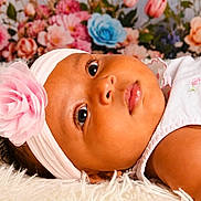 Camilla is registered to the contest to win money with this photo: baby, infant, headband, flower, pink, white_dress, curly_hair, fluffy_rug, portrait, closeup, face, large_eyes, flower_background, soft_texture, cute, child, indoors, resting, beautiful, adorable