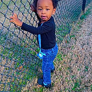 Mosiah is registered to the contest to win money with this photo: afro_hair, black_shirt, blue_jeans, casual_clothing, chain_link_fence, child, crocs, curious, daylight, fence, field, grass, holding, nature, outdoor, pacifier, person, standing, toddler, young_child