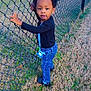 afro_hair, black_shirt, blue_jeans, casual_clothing, chain_link_fence, child, crocs, curious, daylight, fence, field, grass, holding, nature, outdoor, pacifier, person, standing, toddler, young_child